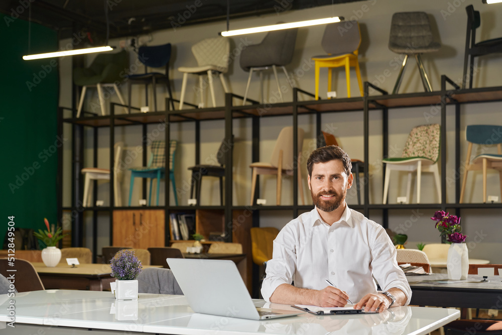 Smiling manager sitting at table with laptop, while working remotely. Front view of confident retailer looking at camera, while writing against rack with chairs on background. Concept of workspace. 