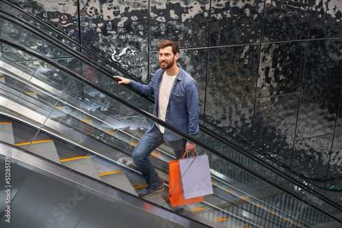 Handsome bearded male holding shopping bags, while lifting up on escalator of shopping center. View from above of smiling man in casual outfit standing on moving staircase inside. Concept of shopping.