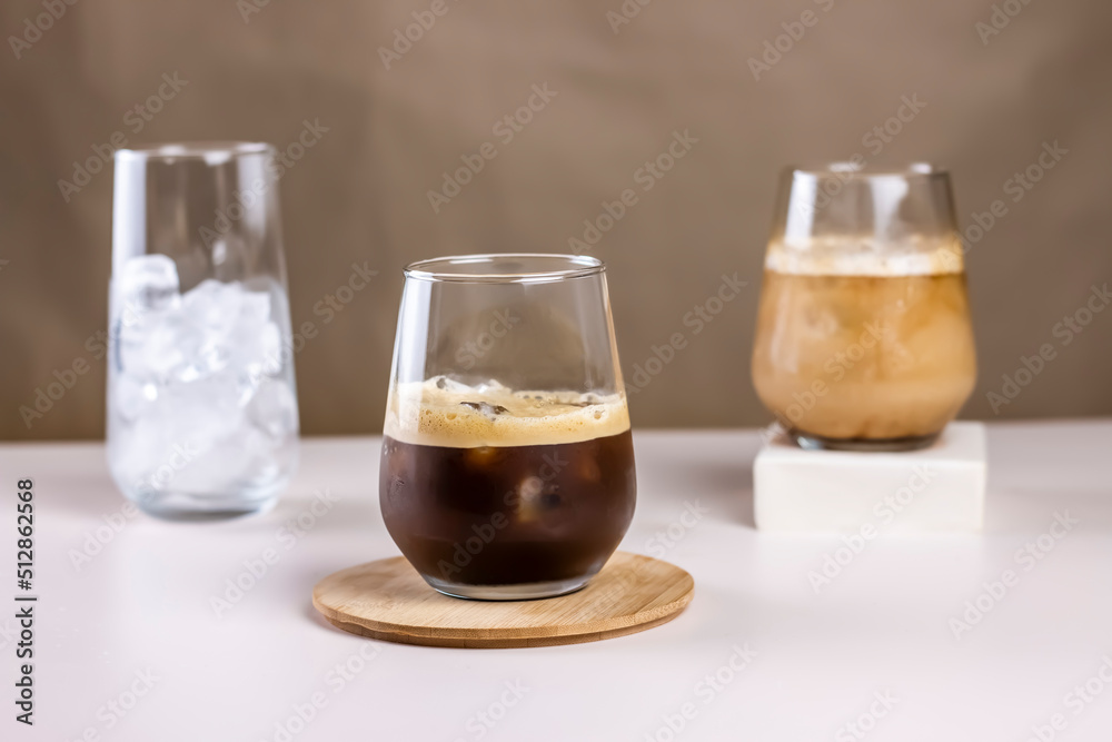 Cold coffee summer drink concept. Different size transparent glasses with ice cube and iced coffee on pastel brown background. Cold coffee summer drink concept. Different size transparent glasses with ice cube and iced coffee on pastel brown background.