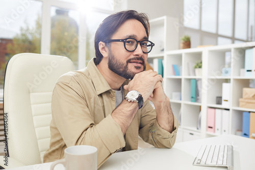 Portrait of content dreamy young Asian designer with beard sitting at desk in office and leaning head on hands