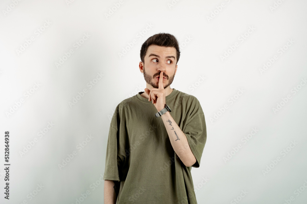 Caucasian young man wearing t-shirt posing isolated over white background makes shush gesture, looks surprisingly aside, notices something strange, has dark stubble.
