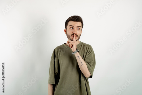 Caucasian young man wearing t-shirt posing isolated over white background makes shush gesture  looks surprisingly aside  notices something strange  has dark stubble.