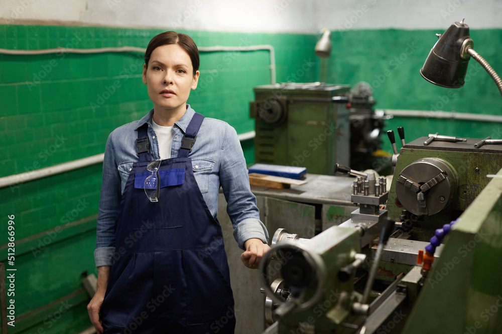 Fototapeta premium Portrait of serious middle-aged woman in overall with hanging safety goggles standing at manual lathe in industry workshop