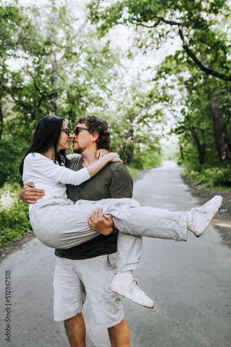 A bearded long-haired stylish man holds his beloved hippie brunette woman in sunglasses in his arms and circles her in nature in a park on the road. Portrait, photo of happy and smiling newlyweds.