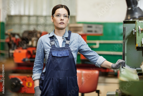 Pensive gritty female manufacturing engineer in blue overalls wearing protective goggles and gloves standing at factory machine