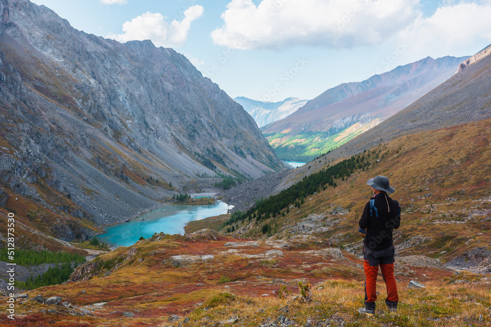 Man enjoys beautiful view of blue alpine lake in colorful autumn valley. Tourist enjoying azure mountain lake scenic view from red hill among multicolor flora. Vivid autumn colors in high mountains.