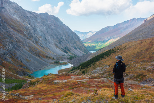Man enjoys beautiful view of blue alpine lake in colorful autumn valley. Tourist enjoying azure mountain lake scenic view from red hill among multicolor flora. Vivid autumn colors in high mountains.