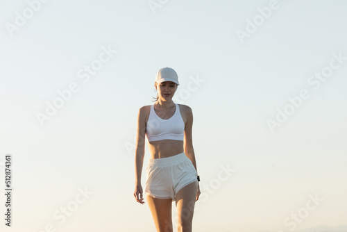 Burnette woman in baseball cap walking in sportive outfit against sky.