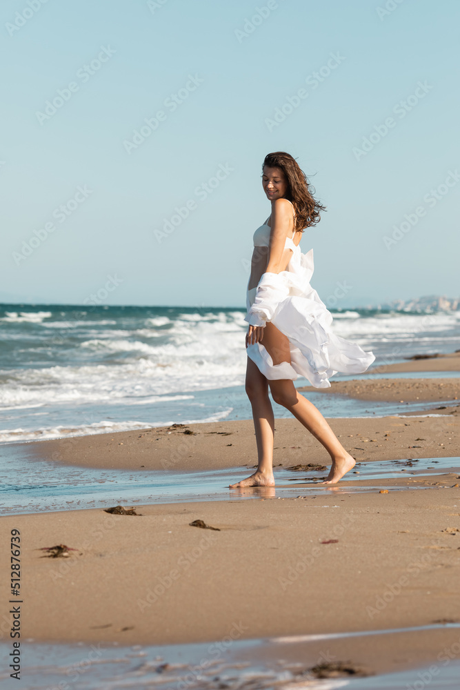 full length of barefoot woman in white shirt and swimwear standing near ocean on wet sand. full length of barefoot woman in white shirt and swimwear standing near ocean on wet sand.