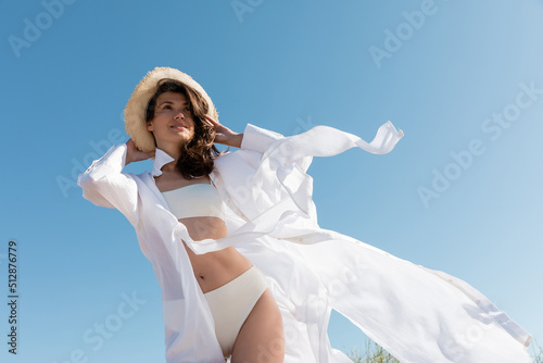 low angle view of young woman in swimsuit and white shirt wearing straw hat against blue sky.