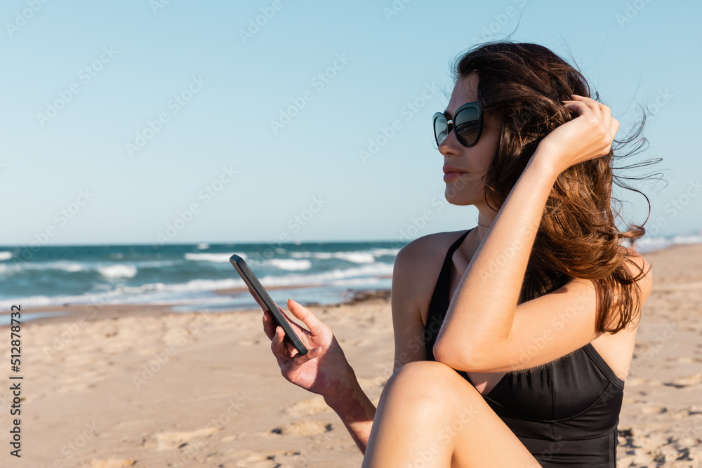 young woman in swimsuit and sunglasses holding smartphone near sea.