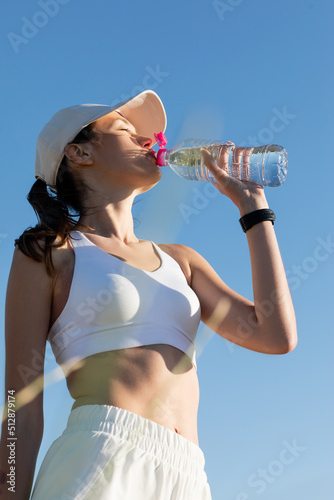 Obraz na plátně low angle view of sportive woman in baseball cap and sports bra drinking refreshing water against blue sky