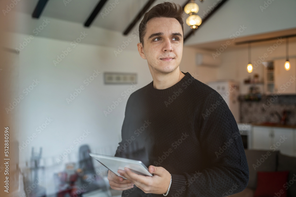 Fototapeta premium One young adult caucasian man standing indoor at home thinking and holding tablet looking to the side - Technology and working from home concept