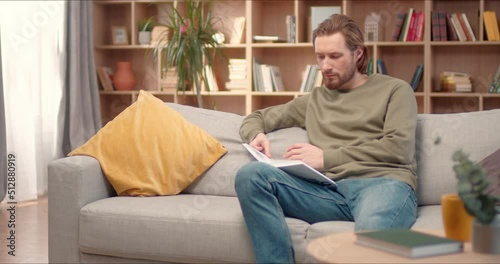 Young handsome caucasian man sitting on couch reading book. Male studying at home selfeducation, covid 19 quarantine. Fiction and self-development literature concept. photo