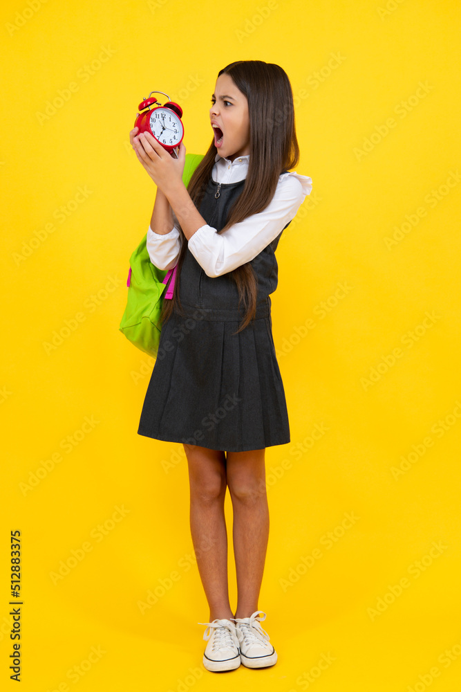 Back to school. Teenager schoolgirl with bag hold clock alarm, time to learn. School children on isolated yellow background. Angry teenager girl, upset and unhappy negative emotion. Back to school. Teenager schoolgirl with bag hold clock alarm, time to learn. School children on isolated yellow background. Angry teenager girl, upset and unhappy negative emotion.