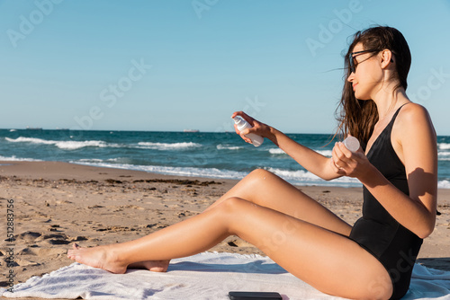 young woman in sunglasses and black swimwear applying sunblock on beach.