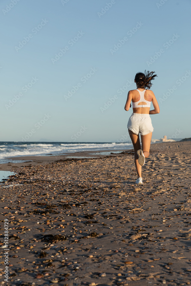back view of sportive woman in white shorts jogging near sea in summer.