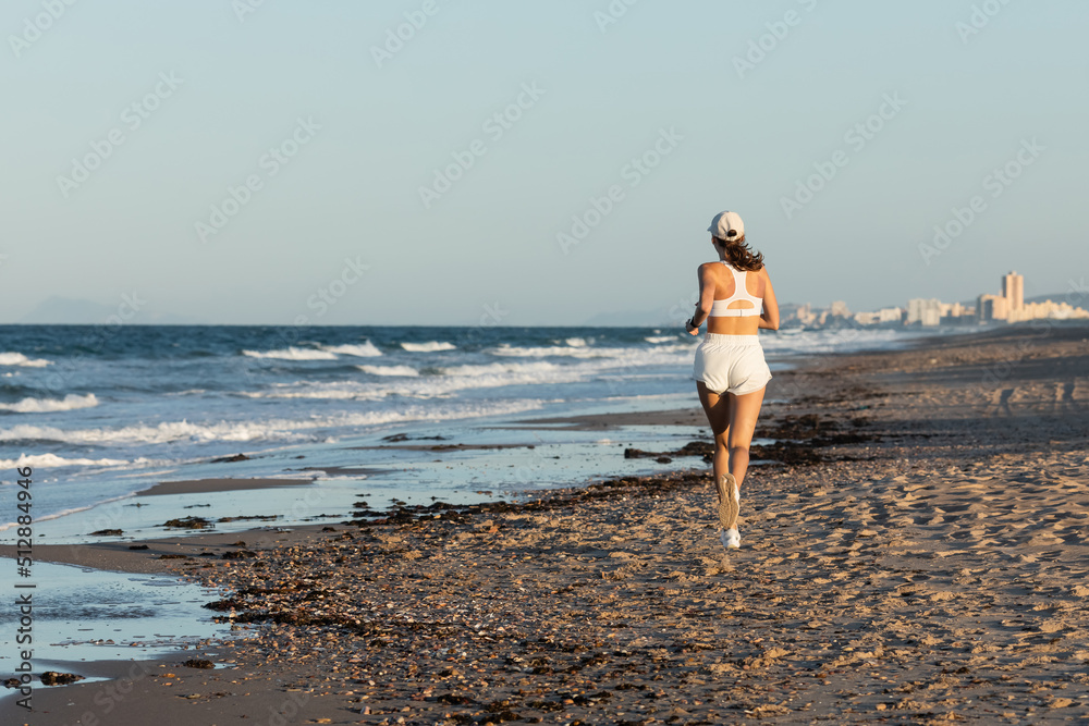 back view of young sportive woman in baseball cap jogging near sea in summer, banner.