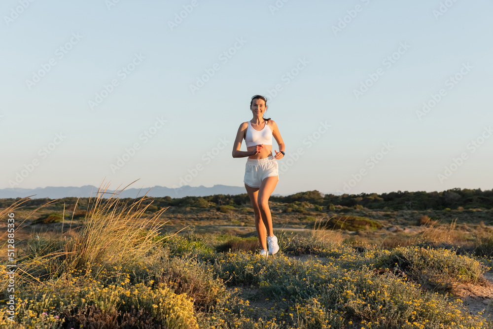 smiling sportswoman in shorts and sports bra listening music and running outside.