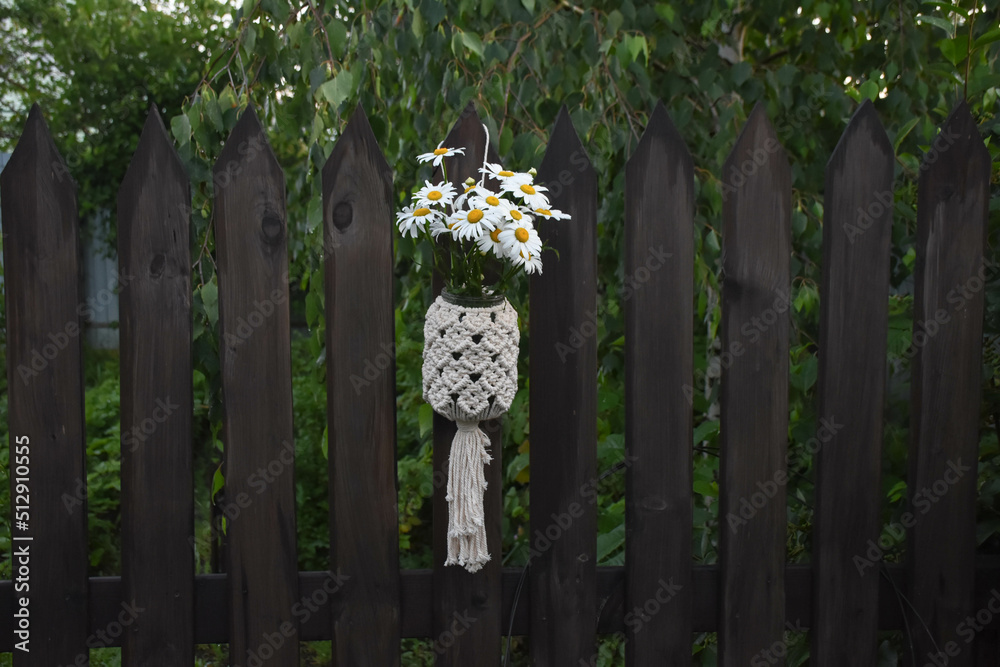 a bouquet of daisies in a macrame vase hanging on a wooden fence