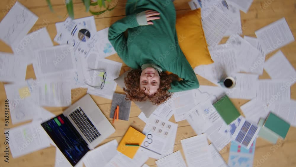 Top down camera roll shot of young female freelancer lying on floor at home surrounded by financial papers and posing for camera with smile Top down camera roll shot of young female freelancer lying on floor at home surrounded by financial papers and posing for camera with smile