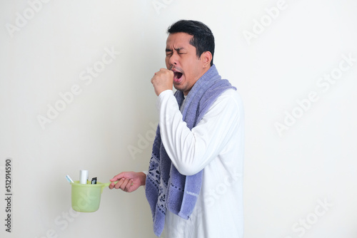 Side view of Adult Asian man yawning while holding shower dipper and bring towel around his neck photo