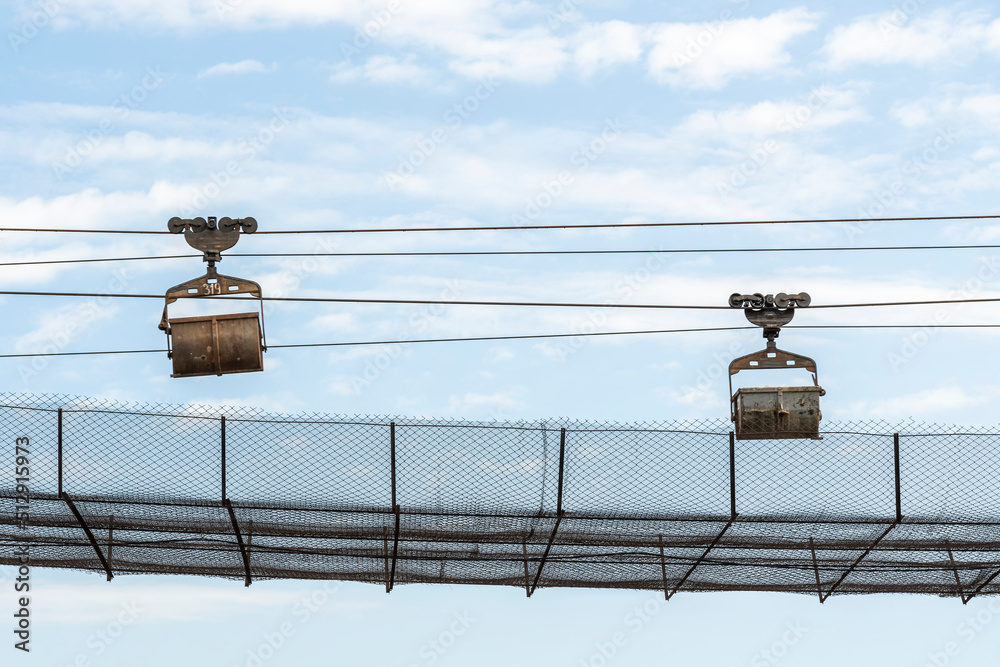 Obraz premium Protected industrial ropeway, on which trolleys move, empty and loaded with raw materials against background of blue summer sky.