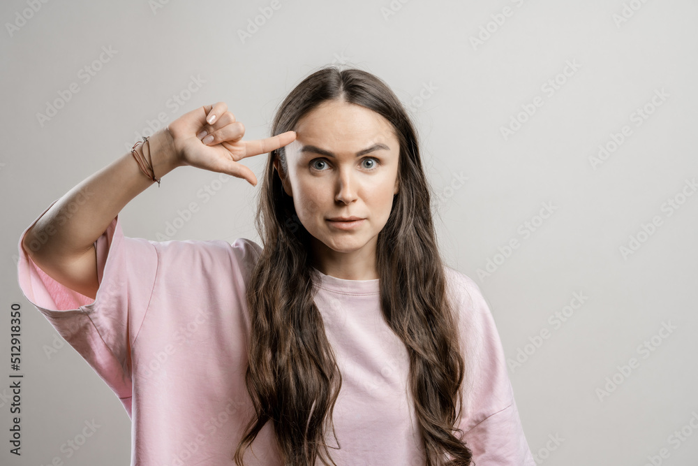 The girl shows her finger at her temple. Brunette girl with long hair on a gray background