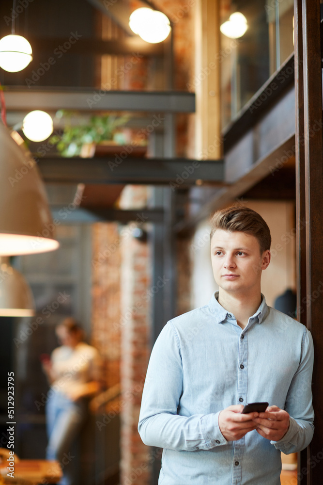 Introspective young handsome restaurant manager in light blue shirt leaning on wall and using smartphone while overseeing work of staff