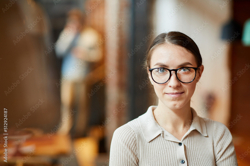 Portrait of smiling attractive young woman in glasses wearing casual sweater standing in public establishment Portrait of smiling attractive young woman in glasses wearing casual sweater standing in public establishment