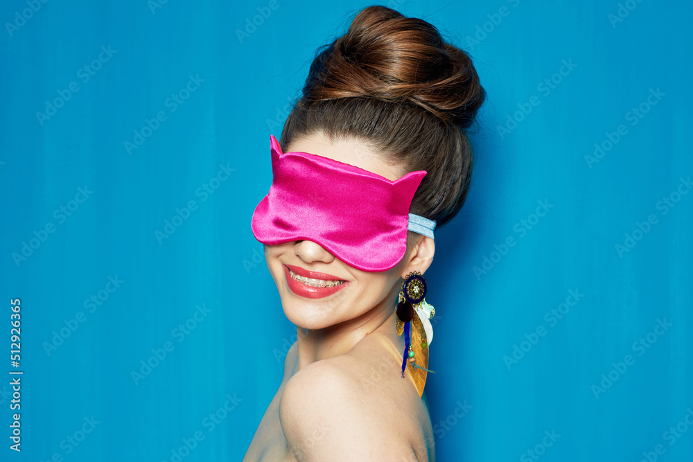 Smiling woman with braces, bare shoulders, pink sleep mask and bun hairstyle isolated on blue wall background.