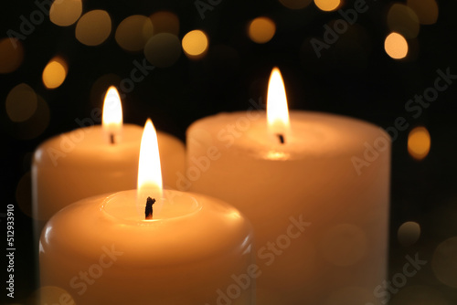 Wax candles burning on black background with blurred lights, closeup. Bokeh effect