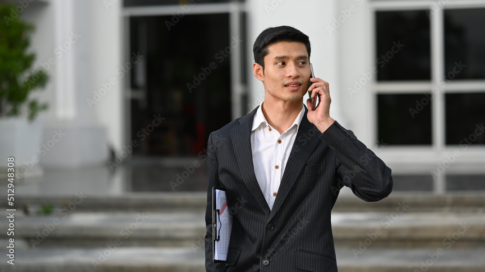 Handsome asian businessman walking outside modern office building and talking with business partner on mobile phone Handsome asian businessman walking outside modern office building and talking with business partner on mobile phone