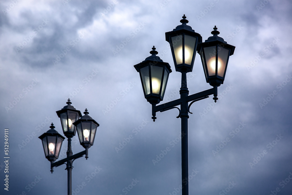 triple lantern 3 head with transparent glass panels and electric bulb, iron street lighting glowing in evening against blue cloudy sky, retro style cast iron lamp pole.