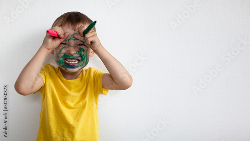 A 4-year-old boy painted his face with a marker. The boy stands against a white wall, holding markers in his hands and portrays a monster with horns. Banner with space for text