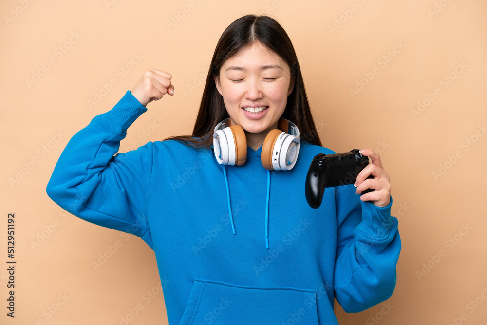 Young Chinese man playing with a video game controller isolated on beige background doing strong gesture