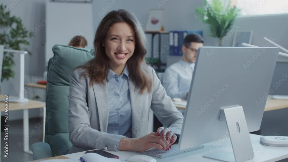 Smiling happy young businesswoman looks at camera working on laptop computer in office. On background people working Smiling happy young businesswoman looks at camera working on laptop computer in office. On background people working