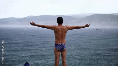 Happy adult male swimmer standing in the pouring rain, arms wide open, by the ocean after a swim. photo