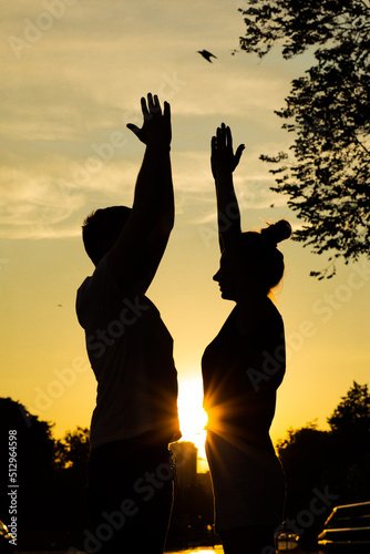 Young couple exercising on street in city from below view, sunset background. Darkened silhouettes of training man and woman looking and standing opposite each other with raising hands