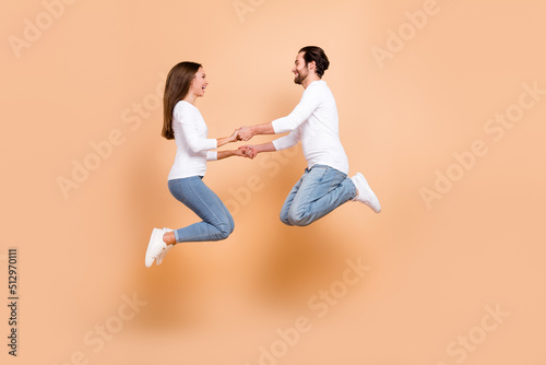 Full length body size view of two attractive cheery partners jumping holding hand isolated over beige pastel color background