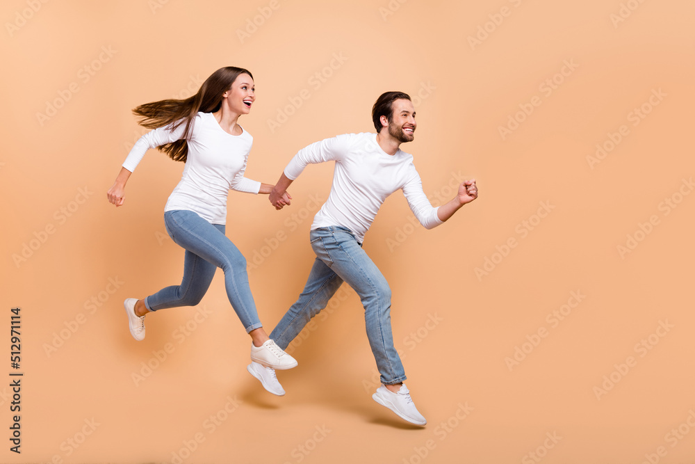 Full length body size view of attractive trendy cheerful partners jumping motion run isolated over beige pastel color background Full length body size view of attractive trendy cheerful partners jumping motion run isolated over beige pastel color background