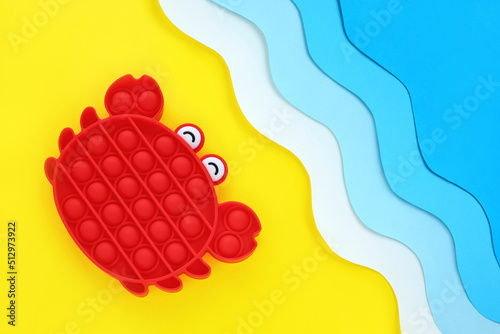 Red crab popular antistress sensory pop it toy on paper cut out seashore diagonal background, yellow sand and blue waves. Relaxation and fun on vacation for the whole family. Summer holidays concept.