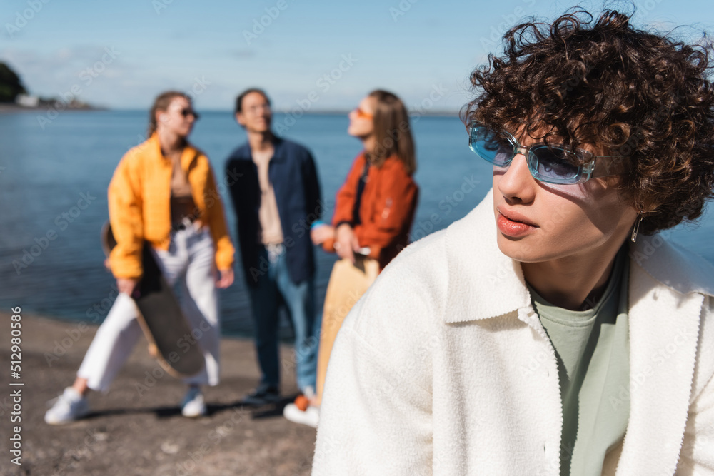 young man in trendy eyeglasses looking away near friends on blurred background.