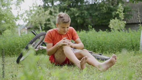 A young handsome blond boy in orange clothes sits on the ground in a green park by a summer house and cries because he fell off a bicycle and injured his knee. Agony of pain due to trauma to the skin.
