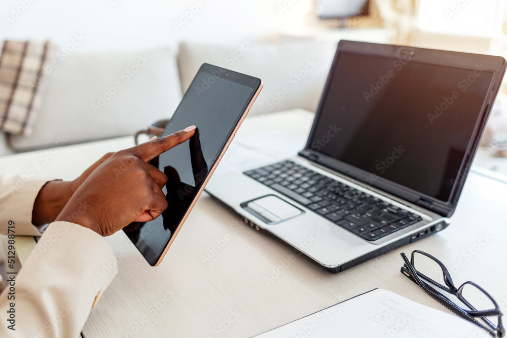Cheerful African American woman, sitting on her desk, smiling and using her laptop. She is typing something. Woman working with tablet computer in the living room during the day. Cheerful African American woman, sitting on her desk, smiling and using her laptop. She is typing something. Woman working with tablet computer in the living room during the day.