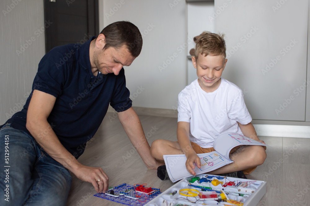 A cute boy in a white T-shirt collects an electrical designer with his dad in the room A cute boy in a white T-shirt collects an electrical designer with his dad in the room