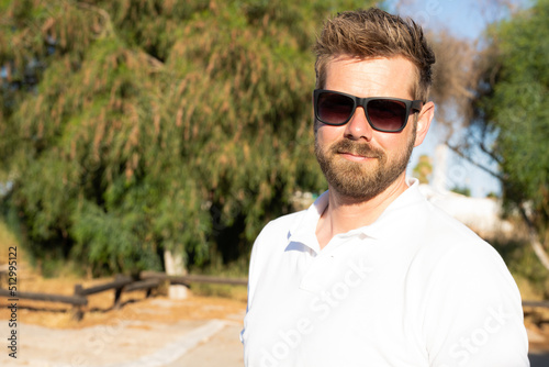 Outdoor shot of beautiful bearded young man in sunglasses posing over city garden on warm sunny day, wearing white polo shirt
