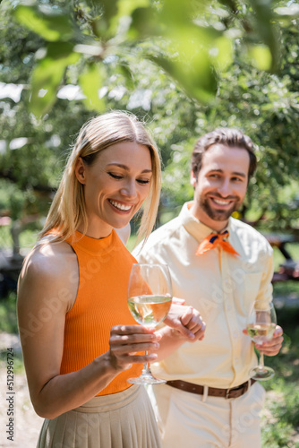Cheerful woman holding glass of wine near blurred boyfriend in summer park.