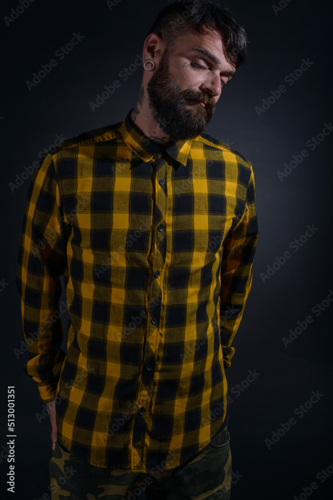 Attractive man with tattoos posing dressed in yellow and black checkered shirt on a black background Attractive man with tattoos posing dressed in yellow and black checkered shirt on a black background