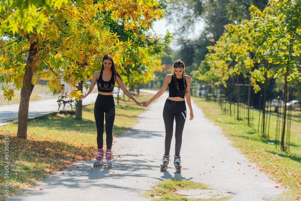 Obraz premium Beautiful sweet girlfriends riding on roller skates and holding hands on the street.